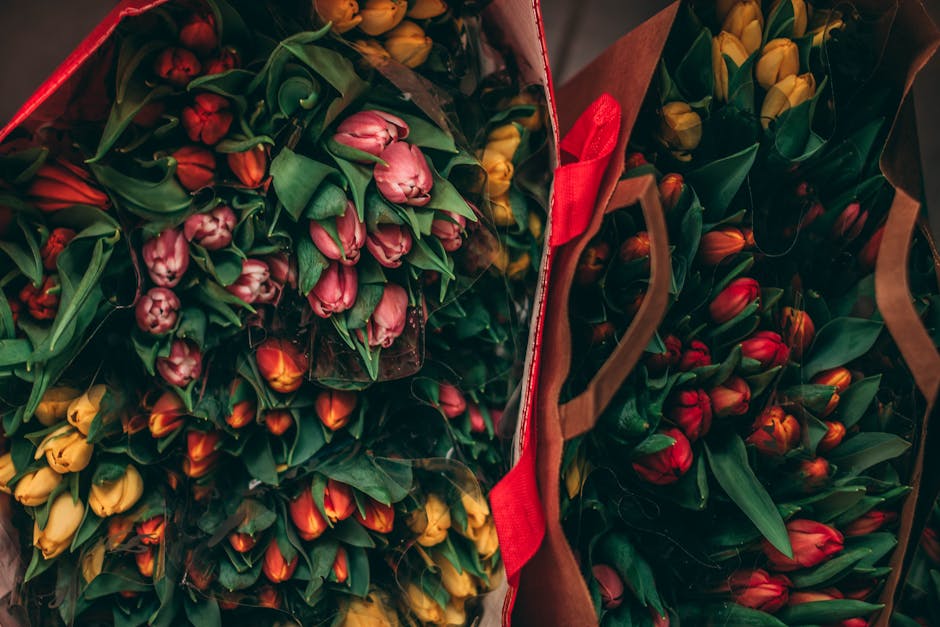 A collection of vibrant tulip bouquets featuring shades of pink, yellow, orange, and red, arranged in protective packaging with green leaves and stems visible, set inside open gift bags with handles.